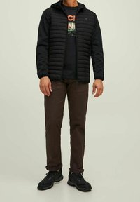 Chaqueta acolchada negra con capucha, combinada con una camiseta gráfica oscura, pantalones marrones y zapatos de senderismo negros. Diseño simple y funcional.