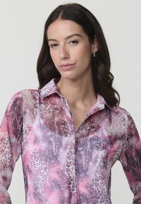 Blusa transparente con un estampado animal en tonos rosa y púrpura, con un sutil patrón de piel de serpiente. Cuello clásico y cierre de botones en la parte delantera.