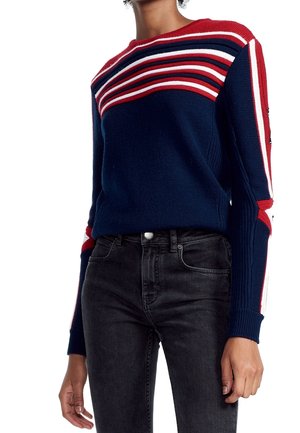 Osoba nosící tmavě modrý svetr s proužky v červené, bílé a modré barvě na ramenou a rukávech, spojený s černými džínami.
