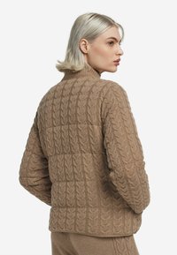 Brun kabelstickad tröja med hög krage, strukturerat mönster och avslappnad passform. Figursydd vid midjan med ribbad kant.