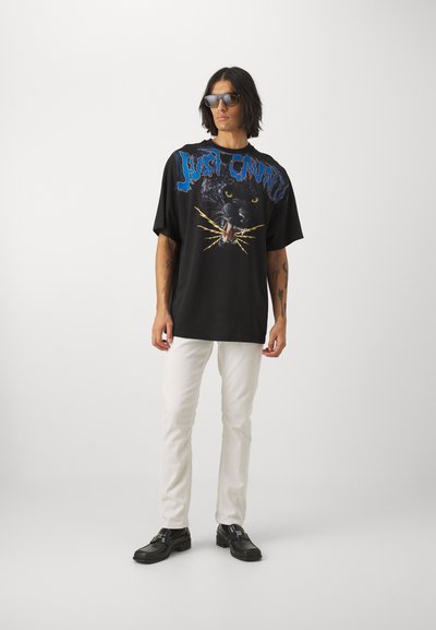 T-shirt nera oversize con una stampa grafica di un pantera con accenti blu e dorati. Abbinata a jeans slim-fit bianchi e scarpe nere.