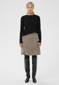 Schwarzer Pullover mit langen Ärmeln, beigefarbener Wildlederrock bis zum Knie und kniehohe schwarze Lederstiefel, präsentiert an einem Model vor einfarbigem Hintergrund.