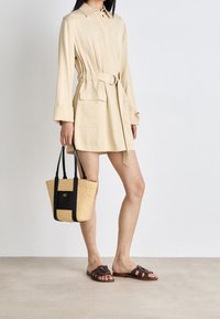 Robe chemise beige à ceinture avec deux poches avant, assortie à des sandales plates brunes et à un sac fourre-tout tissé beige et noir. Texture de tissu lisse.