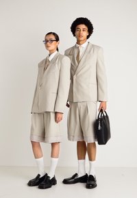 Duas modelos vestindo blazers oversized bejes, calções largos combinando, meias brancas e mocassins pretos; uma delas segura uma bolsa preta.