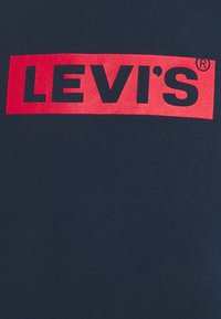 T-shirt en coton bleu marine avec un grand logo rectangulaire rouge portant l'inscription "LEVI'S" en lettres noires en gras. Texture lisse.