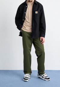 Sort skjorte med knapper over beige turtleneck, olivengrønne bukser og sort-hvide sneakers med mønster. Simpelt design og neutrale farver.