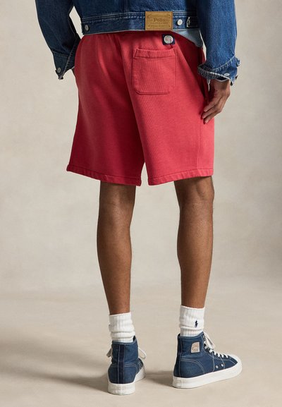 Homme portant un short rouge avec une poche arrière, une veste en jean, des chaussettes blanches et des baskets montantes bleues, debout sur un fond neutre.