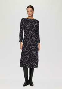 Schwarzes Midi-Kleid mit langen Ärmeln, das mit weißen abstrakten Herzmustern versehen ist. Es hat einen runden Ausschnitt und eine taillierte Taille. Kombiniert mit schwarzen knielangen Stiefeln.
