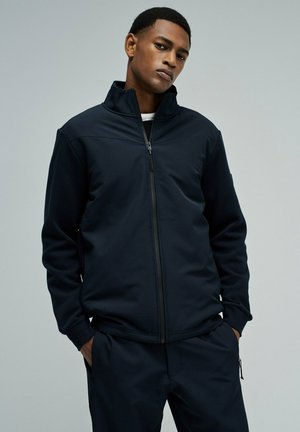 Jeune homme portant une veste noire zippée et un pantalon assorti, debout les mains dans les poches devant un fond gris clair uni.