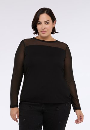 Mujer vistiendo una blusa negra de manga larga con parte superior del pecho y mangas transparentes, combinada con pantalones negros que presentan sutiles adornos.