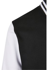 Svart og hvit hettegenser med svart øvre halvdel og hvit nedre halvdel med tekstur. Stripete krage i hvitt, svart og gull.