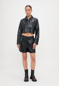 Čierna skrátená bunda z umelého kože s predným zipsom a golierom, v kombinácii s čiernymi kraťasmi z umelého kože. Model nosí čierne členkové topánky.