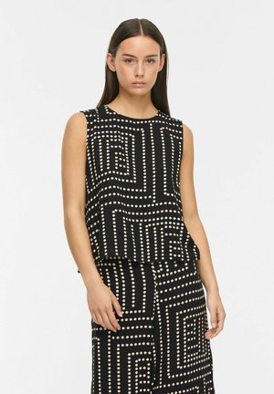 Femme portant un haut noir sans manches et une jupe assortie avec des motifs carrés géométriques beiges à pois, debout sur un fond blanc.