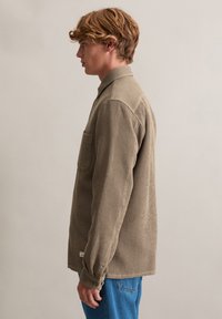 Lichtbruin shirt met wafeltextuur, lange mouwen, een borstzak en knoopmanchetten, gecombineerd met blauwe spijkerbroek.