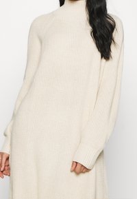 Abito maglione di colore crema con collo alto, polsini a costine e vestibilità rilassata. La texture è morbida e presenta un'intrecciatura morbida e ariosa.