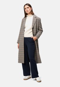 Langternet jakke i brun, beige og sort, over en cremefarvet sweater, kombineret med navy vidde bukser og hvide sneakers.