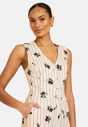 Nainen, jolla on päällään hihaton, napitettu beige liivi mustilla kukka- ja pystysuorilla raidallisilla kuvioilla, käsi lepää sopivien housujen taskussa.