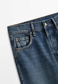 Jean en denim bleu montrant la ceinture, les passants de ceinture, le bouton avant et la petite poche à monnaie sur le côté droit.