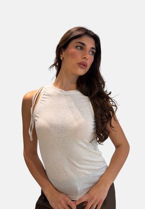 Donna con lunghi capelli scuri e ondulati, indossa una canotta bianca senza maniche con lacci sulle spalle, guarda leggermente verso destra su uno sfondo uniforme.