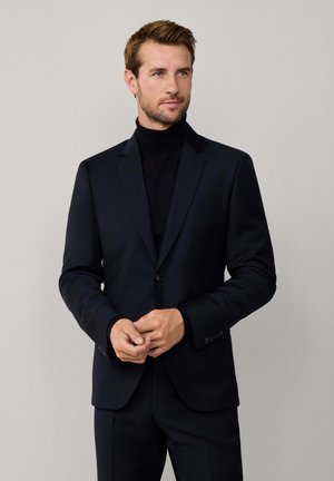 Hombre con traje negro y cuello vuelto parado frente a un fondo gris claro liso, con las manos juntas delante, mirando hacia un lado.