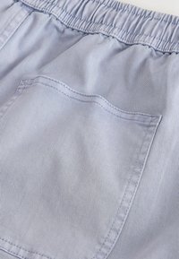 Détail d'un pantalon gris clair à taille élastique montrant la texture du tissu et une poche arrière cousue avec des coutures visibles.