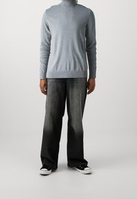 Hellgrauer Rollkragenpullover mit geripptem Saum, kombiniert mit dunkelgrauer Denim-Jeans, leicht ausgewaschen; getragen mit schwarzen Low-Top-Sneakers.