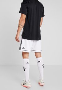 Camisa atlética negra con rayas blancas en los hombros, combinada con pantalones cortos blancos que presentan detalles en negro. Medias blancas y botines completan el look.