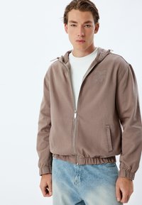 Bruine zip-up jack met capuchon, elastische manchetten en zoom, met een geborduurd logo op de borst. Draagt over een wit shirt en blauwe jeans.