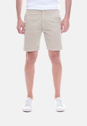 Homme portant un short beige avec ourlet roulé, un t-shirt blanc et des baskets blanches, debout devant un fond uni.