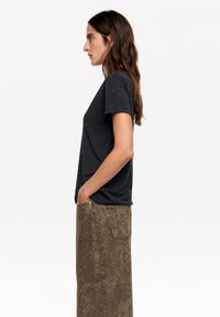 Czarny krótki T-shirt z małymi dekoracyjnymi akcentami. Noszony z długą, wzorzystą brązową spódnicą. Modelka stoi bokiem.