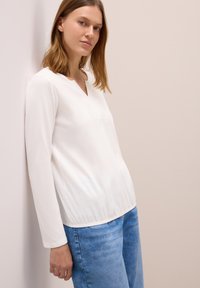 Weißes Langarmoberteil mit V-Ausschnitt und glatter Textur, kombiniert mit hellblauen Jeans, vor einem neutralen Hintergrund.