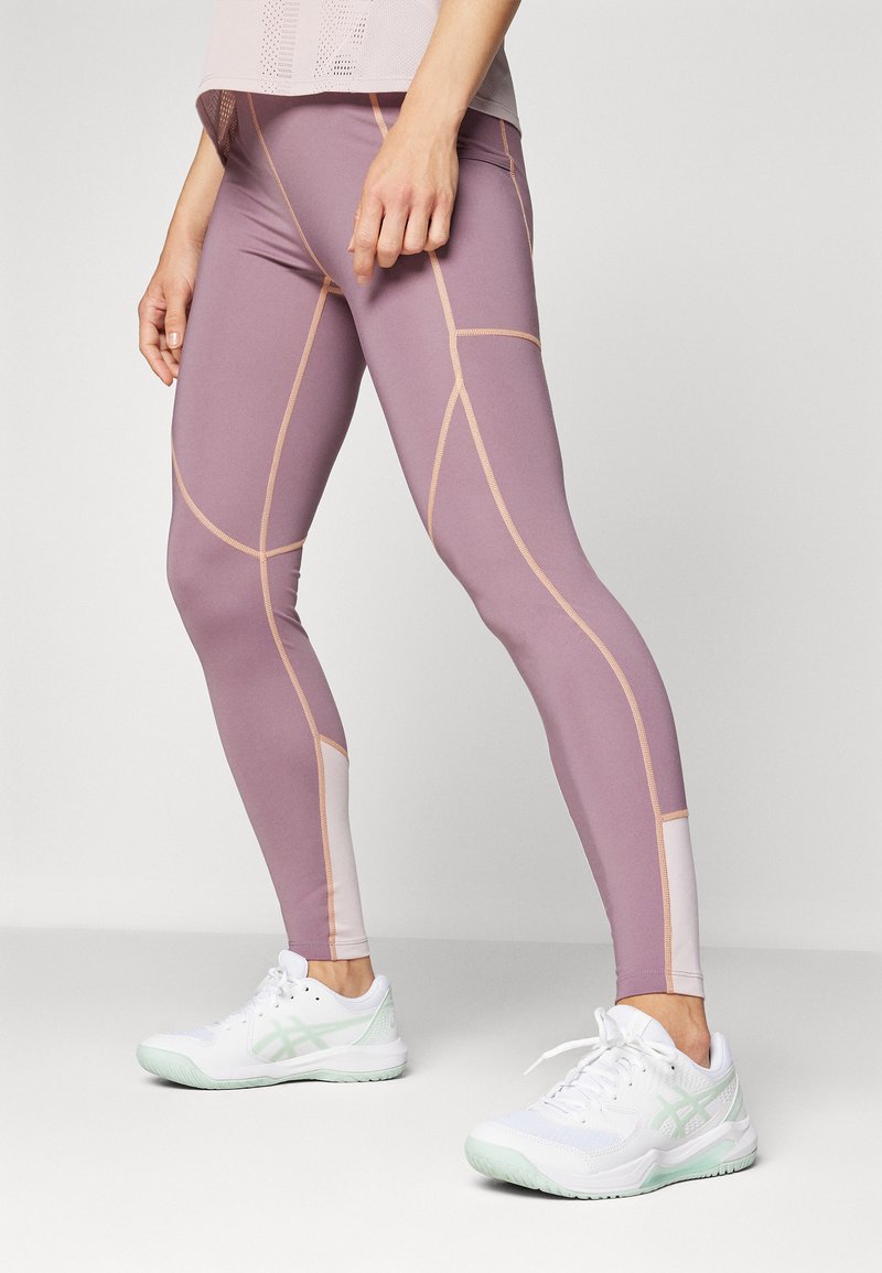 Leggings en lavande doux avec des coutures beiges contrastantes, au design ajusté et avec des panneaux texturés. Associés à des chaussures de sport blanches avec des accents menthe.
