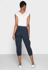 Débardeur blanc côtelé avec des détails en dentelle aux épaules, associé à un pantalon court bleu marine et des baskets blanches avec des accents noirs.