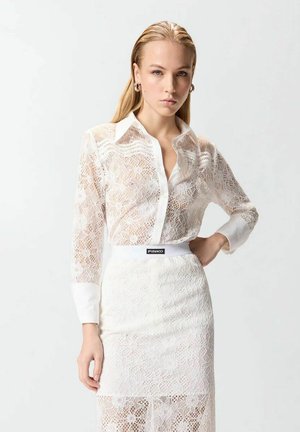 Giovane donna con capelli biondi lisci che indossa una camicetta in pizzo bianco e una gonna in pizzo abbinata, in piedi con una mano sul fianco.