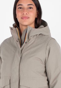 Giacca beige chiaro con cappuccio, dotata di due tasche frontali, chiusura zip e un morbido rivestimento interno con un dettaglio di colore contrastante.