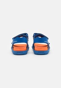 Sandalias amigables para el agua de color azul, fabricadas con material sintético, que cuentan con correas ajustables y una suela naranja, con patrones de agarre texturizados.