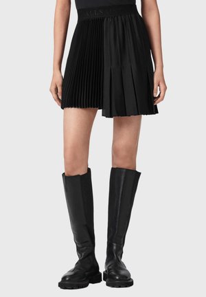 Kvinde iført en sort plisseret mininederdel med et præget linning og høje sorte læderstøvler, stående mod en ensfarvet baggrund.