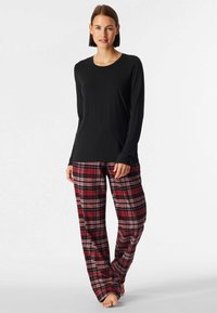 Maglietta nera a maniche lunghe abbinata a pantaloni del pigiama a quadri rossi e neri, con una vestibilità ampia e tessuto morbido. A piedi nudi su uno sfondo semplice.