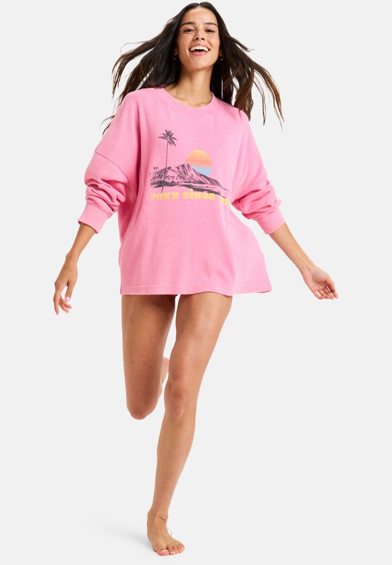 Overdimensjonert rosa genser med grafisk design av palmer og solnedgang. Laget av mykt stoff med lange ermer og avslappet passform.