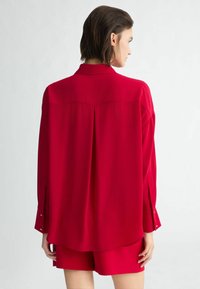 Camicia di seta rossa con piega centrale sul retro, maniche lunghe con polsini abbottonati e colletto classico, mostrata con pantaloni corti coordinati.