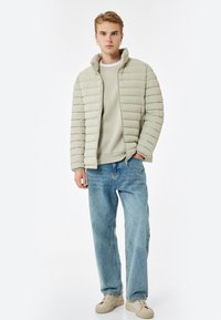 Jasnozielona kurtka puchowa z poziomym pikowaniem, wysoki kołnierz i zamek z przodu, w zestawie z jasnoszarym swetrem i niebieskimi dżinsami.
