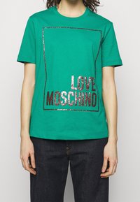 Person som bär en grön kortärmad T-shirt med glänsande mörkrosa texten "LOVE MOSCHINO" inuti en rektangulär kant, i kombination med mörka jeans.