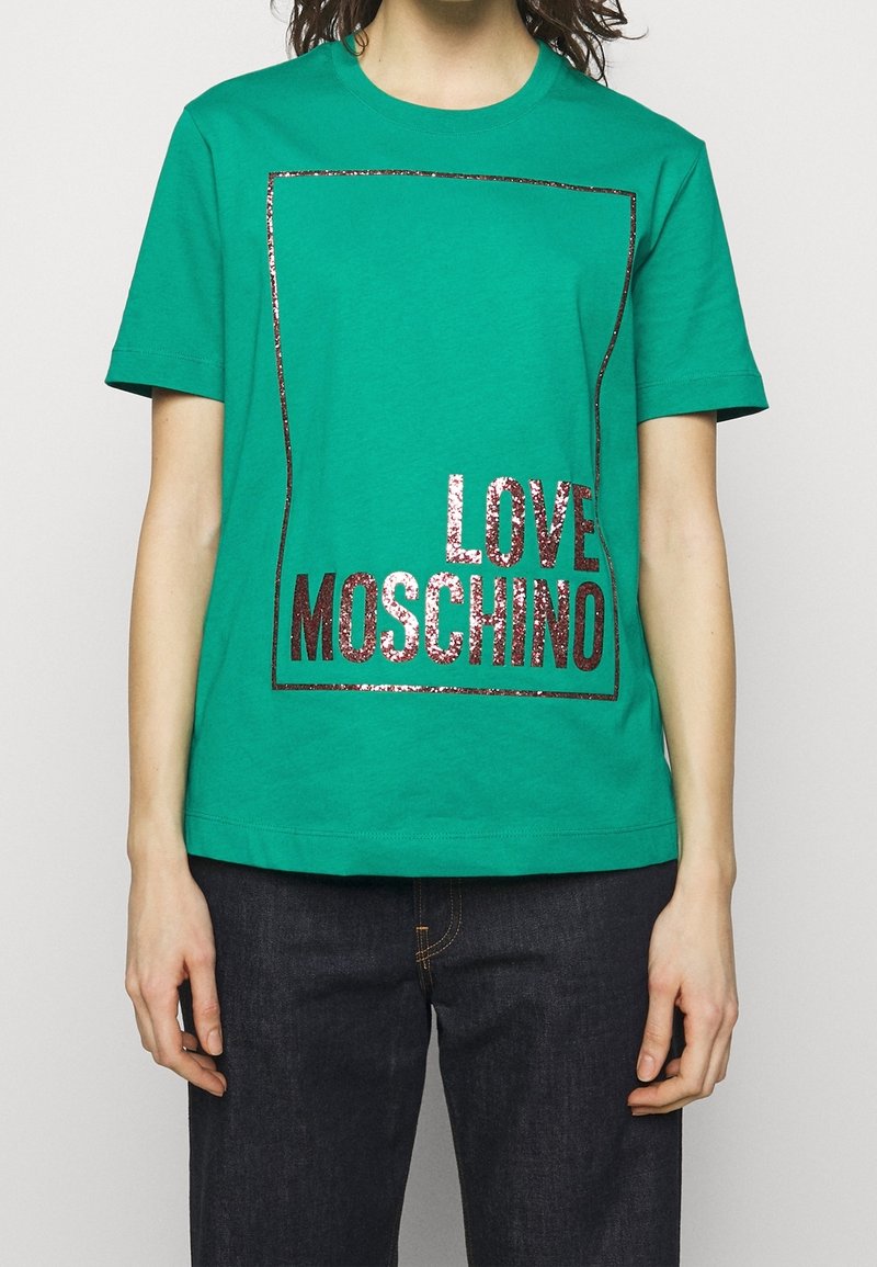 Person som bär en grön kortärmad T-shirt med glänsande mörkrosa texten "LOVE MOSCHINO" inuti en rektangulär kant, i kombination med mörka jeans.