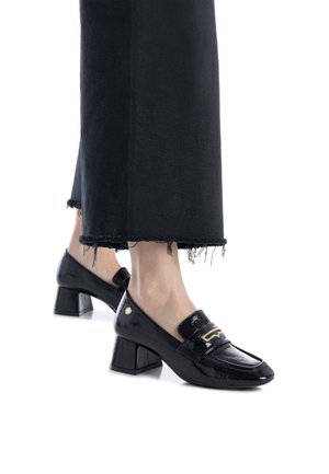 Mocasines negros de charol con tacón cuadrado y una correa de acento dorada, combinados con pantalones de mezclilla negros deshilachados.
