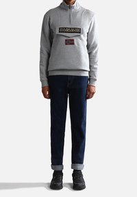 Pullover grigio in pile con zip a metà, dotato di una toppa logo frontale e una bandiera norvegese. Abbinato a jeans scuri e sneaker nere.