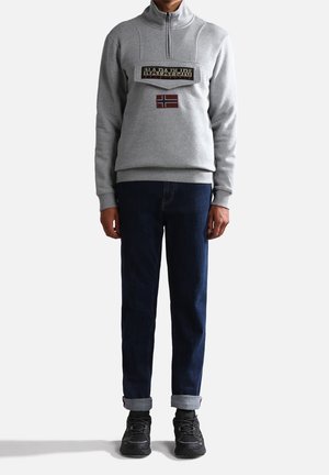 Pullover grigio in pile con zip a metà, dotato di una toppa logo frontale e una bandiera norvegese. Abbinato a jeans scuri e sneaker nere.