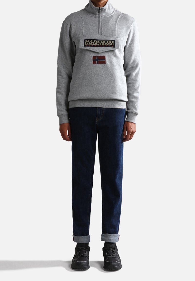 Pullover grigio in pile con zip a metà, dotato di una toppa logo frontale e una bandiera norvegese. Abbinato a jeans scuri e sneaker nere.