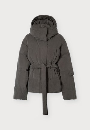 Le mannequin se tient face à l’objectif, vêtu d’une doudoune gris foncé ceinturée, d’un jean noir à jambes larges et de chaussures noires, sur un fond clair uni.