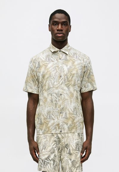 Chemise à manches courtes de couleur beige clair avec un imprimé de feuilles de palmier. Le tissu présente une texture lisse et un col boutonné. Portée avec un short assorti.