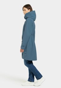 Manteau bleu résistant à l'eau avec une grande capuche, taille cintrée, fermeture éclair à l'avant et deux poches latérales, associé à un jean bleu et des baskets blanches.